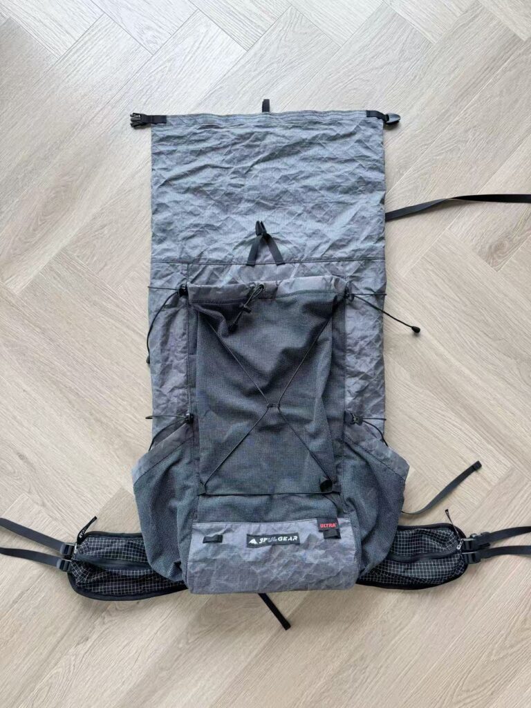 Gray outdoor hiking backpack laid flat on a light wood floor, showing front pocket, bungee cords, and straps central to the bag's design.